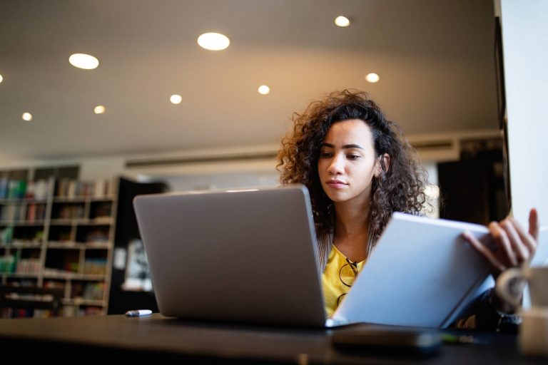 a woman wearing a yellow top working in front of a laptop whilst holding a white folder.