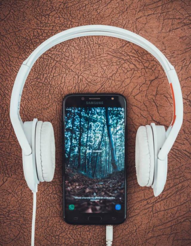 A Samsung smartphone displaying a forest wallpaper with the time "12:37," placed on a brown textured surface. A pair of white wired headphones is wrapped around the phone, forming a circular shape, suggesting a theme of music, relaxation, or digital connectivity.