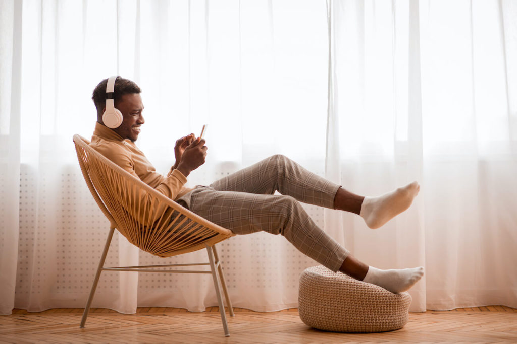 Man sitting in bucket seat with his foot on a pouffe, listening to music through white headphones whilst holding his mobile phone