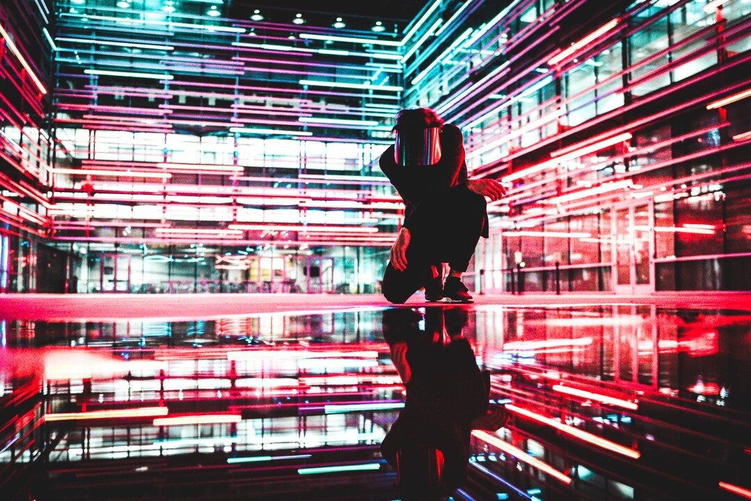 A futuristic, cyberpunk-inspired scene featuring a person crouching in the middle of a high-tech, neon-lit environment. The individual is dressed in dark clothing and wearing a reflective visor, concealing their face. The surrounding space is filled with vibrant red and blue LED light strips, creating a dynamic, digital atmosphere. The glossy floor reflects the neon lights and the person's silhouette, adding depth and symmetry to the composition. The background consists of a glass-walled structure, enhancing the modern, technology-driven aesthetic.