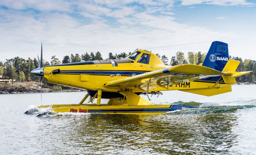 Yellow Saab-branded firefighting plane skimming across a lake, collecting water for aerial fire suppression.