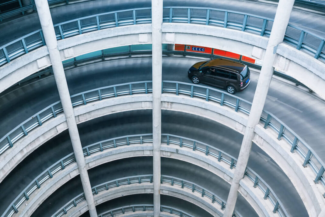 A black car driving on a spiral ramp inside a multi-level circular parking garage. The photo is taken from above, showcasing the repetitive architecture and curves of the structure, highlighting urban infrastructure and transportation themes.