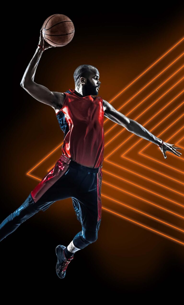 Male basketball player in mid-air wearing a red and black uniform, preparing to dunk the ball with one hand, against a dynamic background of glowing orange lines