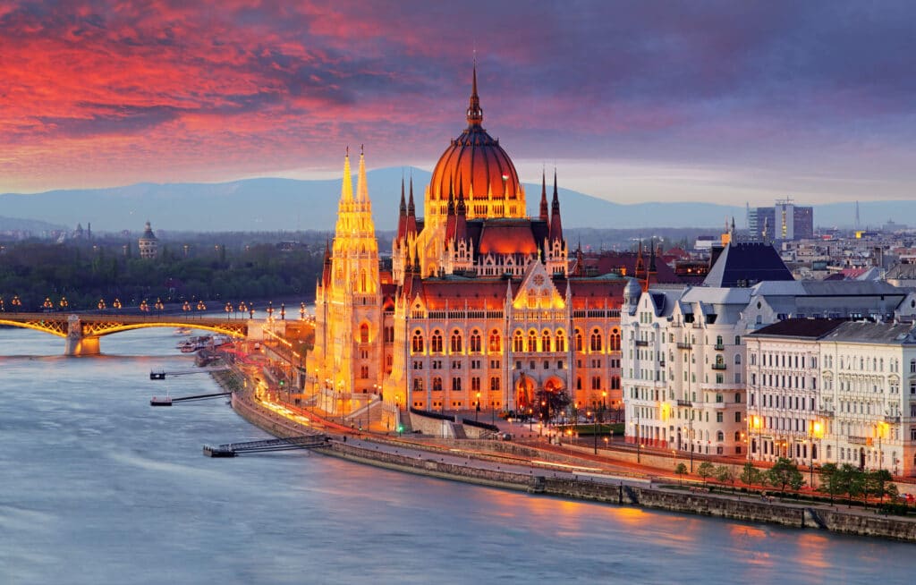 The Hungarian Parliament Building in Budapest illuminated at dusk with the Danube River in the foreground. The sky above is painted with hues of deep orange and pink, contrasting against the building's Gothic Revival architecture. The Margaret Bridge is visible, connecting two parts of the city, with city lights reflecting off the water's surface.