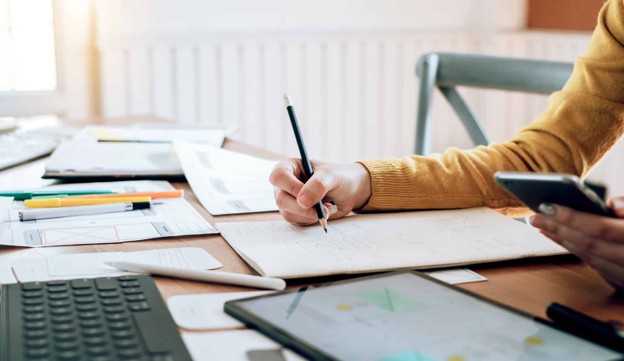 Close-up of a person writing on paper while holding a smartphone, with documents, a tablet, and a keyboard on the desk