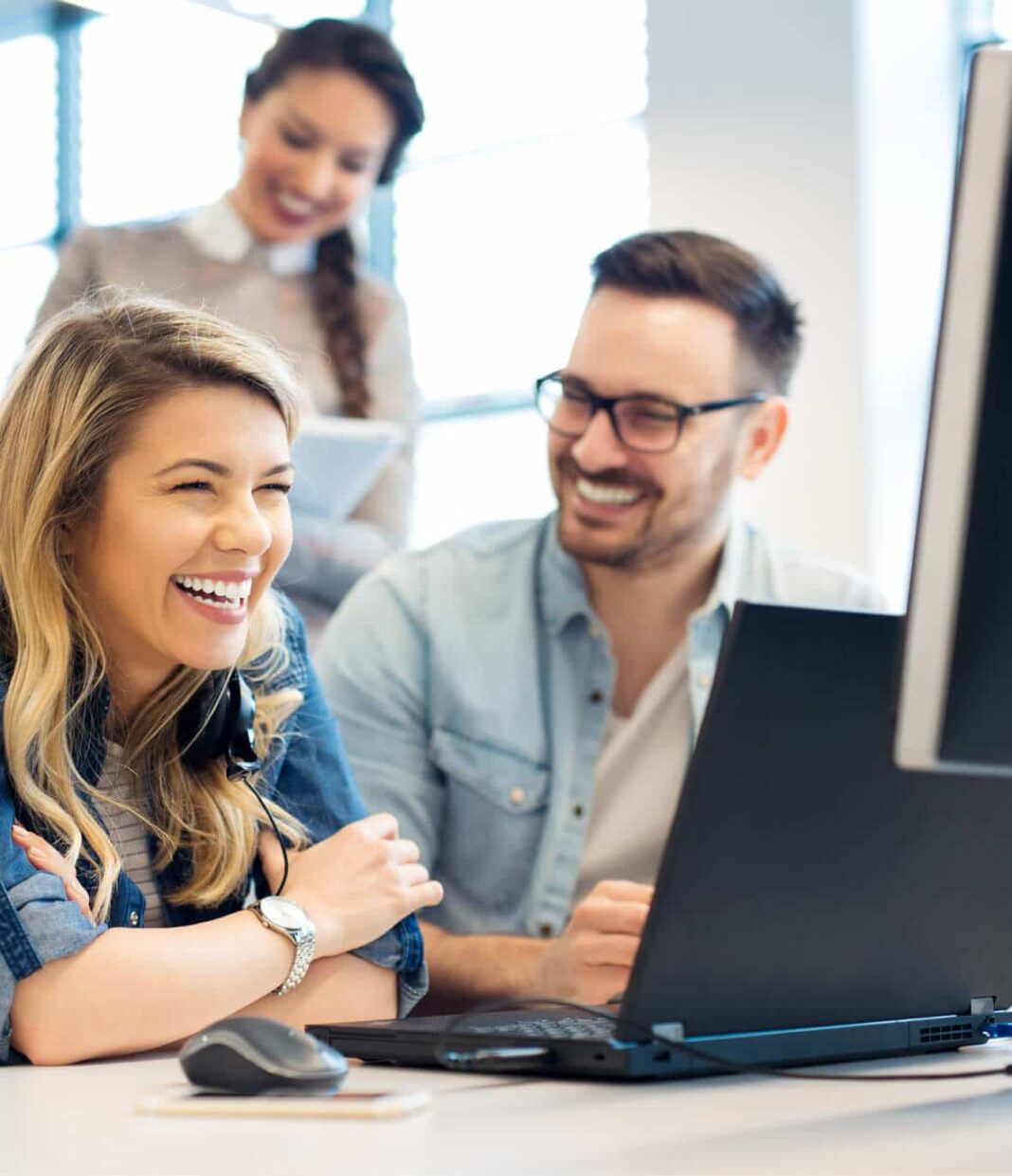Three colleagues smiling and collaborating around a laptop in a bright, modern office setting.