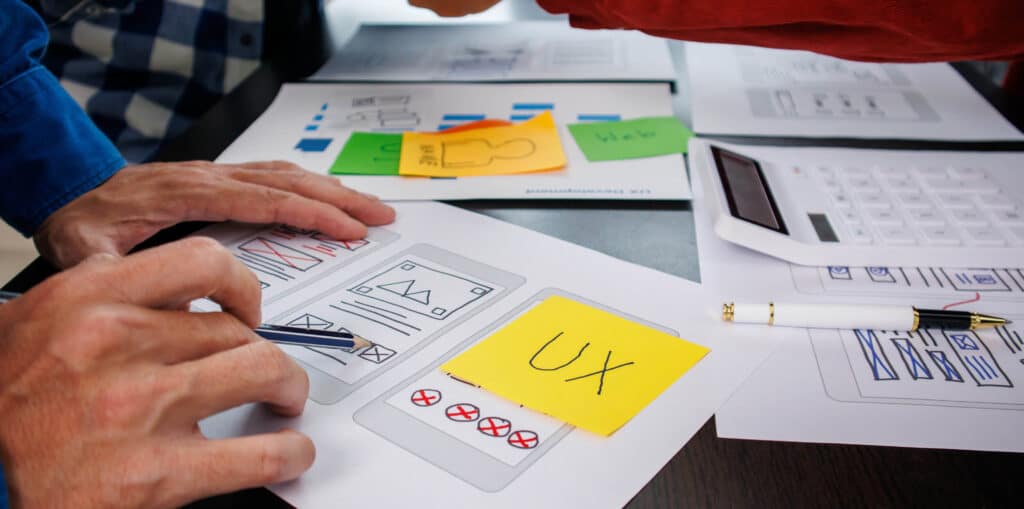 "Hands working on a UX design layout with wireframe sketches and colorful sticky notes on a desk."