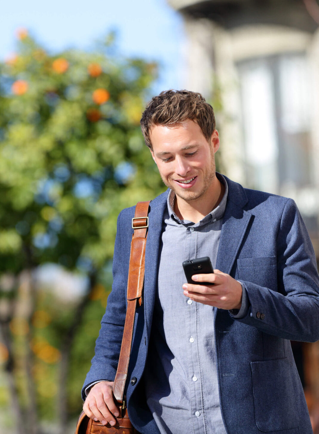 A young man dressed in a smart-casual blazer and shirt, smiling while looking at his smartphone. He carries a brown leather shoulder bag and is walking outdoors on a sunny day, with green trees and blurred buildings in the background, suggesting a relaxed urban setting.