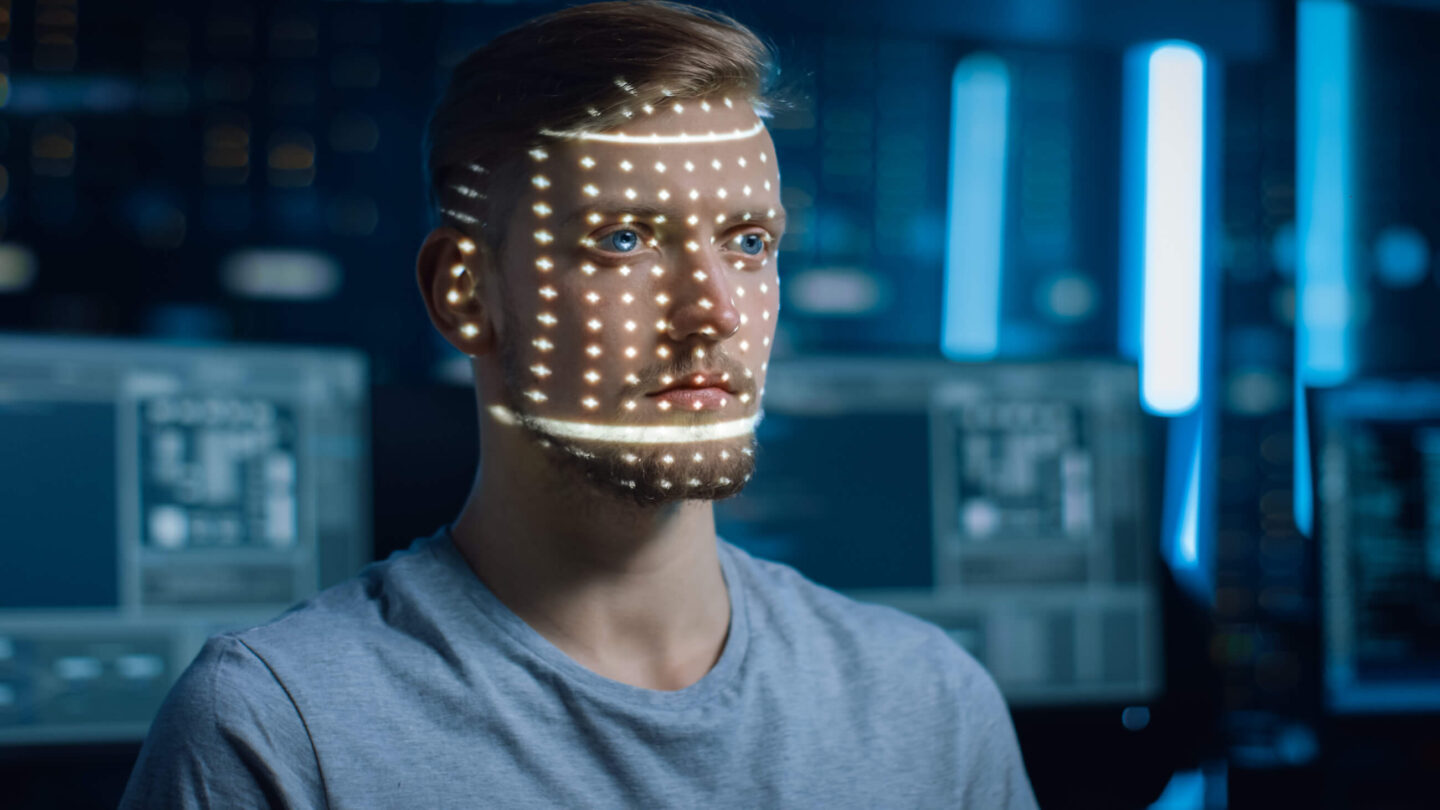 A young man with a beard and blue eyes undergoing facial recognition scanning. A grid of illuminated dots is projected onto his face, analyzing his features in a high-tech environment with multiple computer screens in the background.