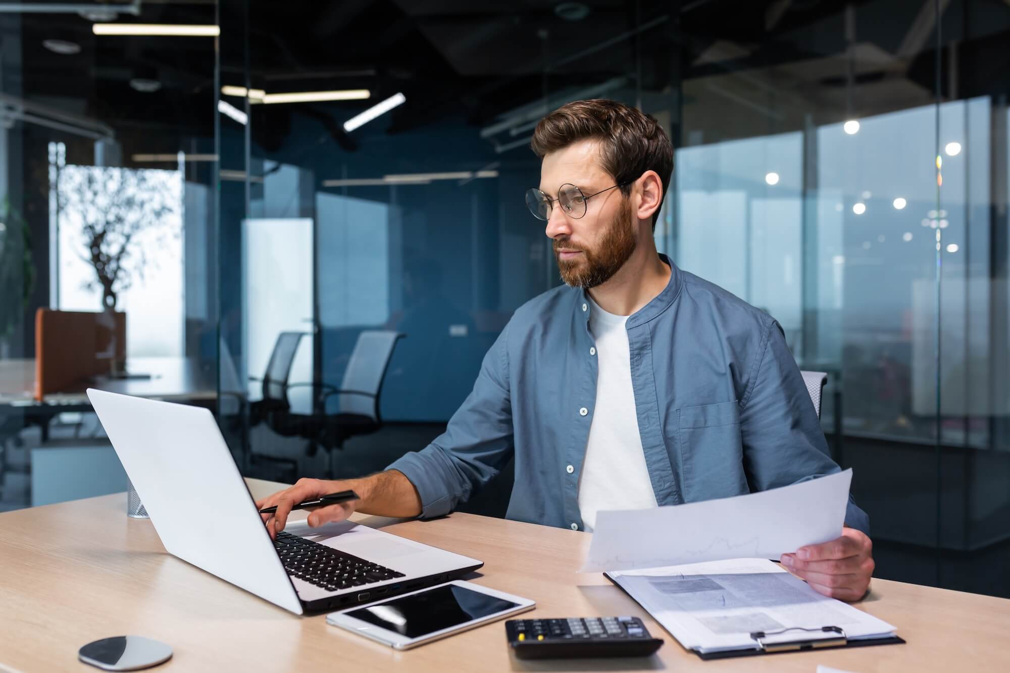 Man in blue overshirt sat at desk with laptop and papers