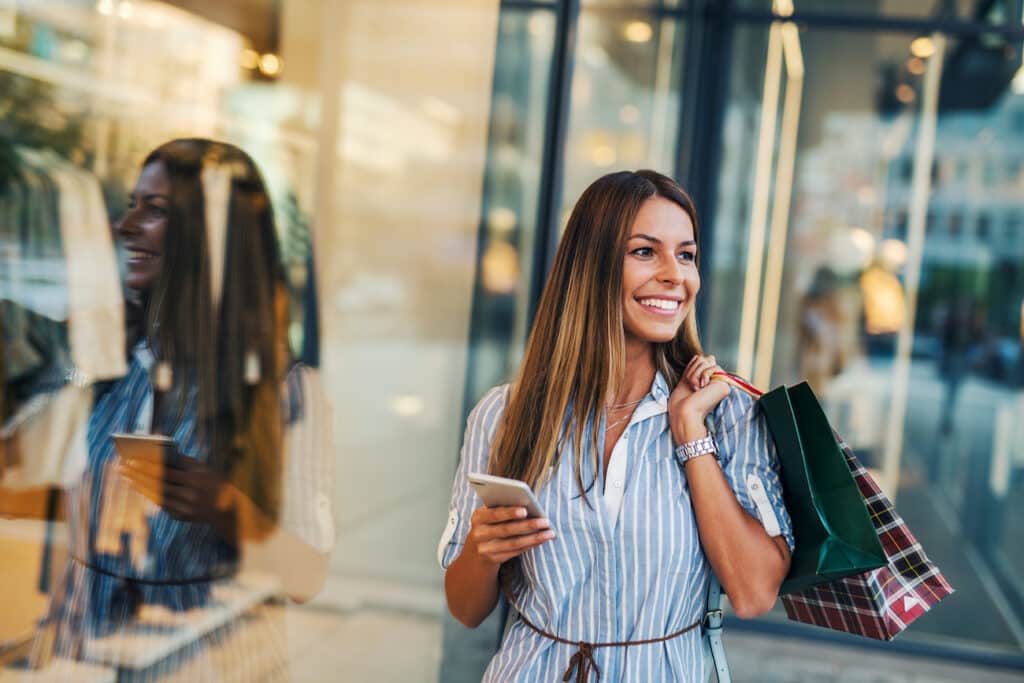 A woman holding a shopping bag over her shoulder with a phone in her other hand stood in front on store window.