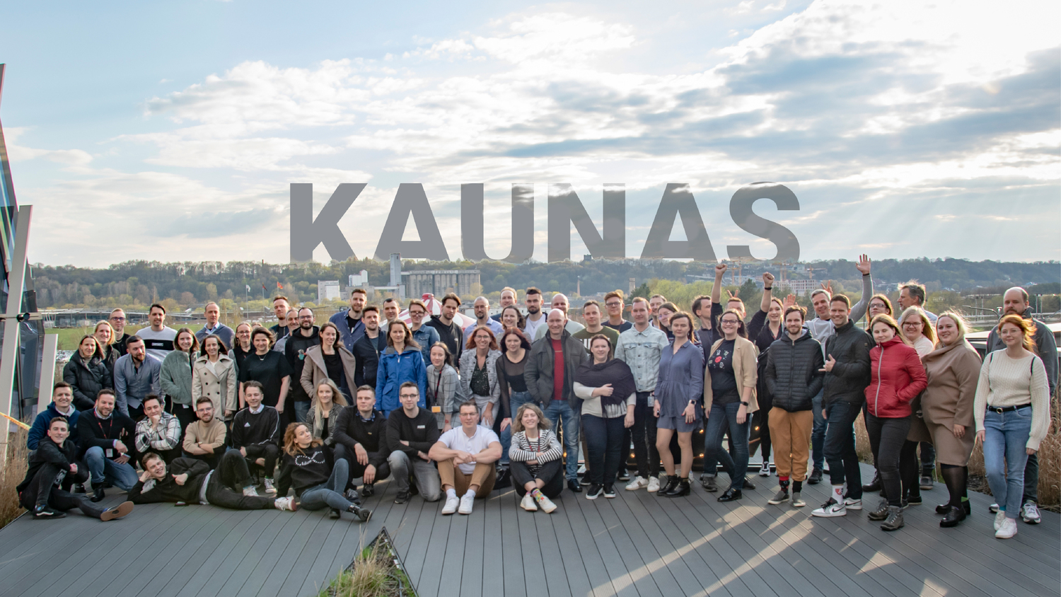 Large group of people smiling and posing outdoors on a terrace with the word “KAUNAS” in large letters in the background.