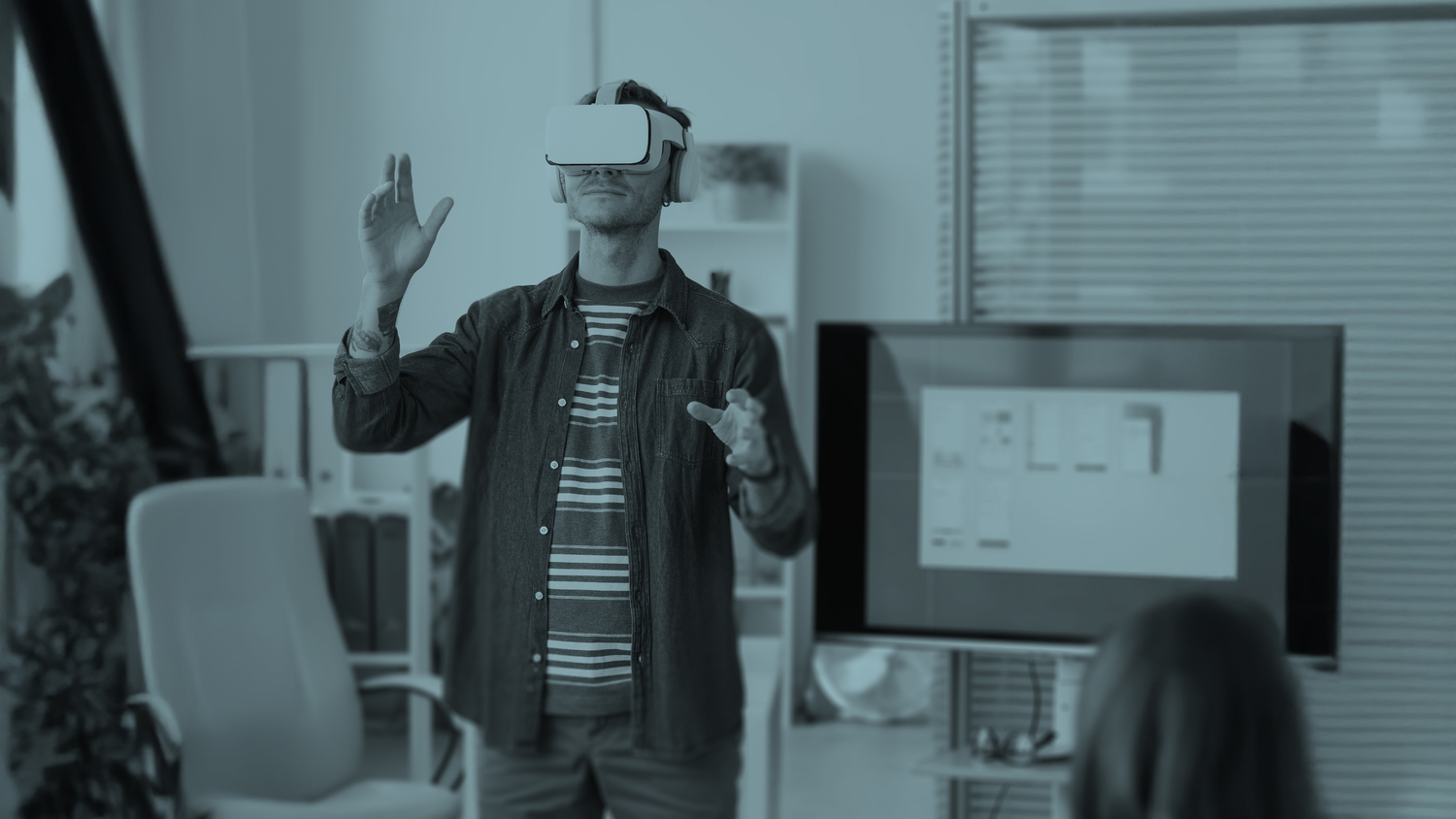 Man wearing a virtual reality headset with arms raised, interacting with a virtual environment in a modern office setting.