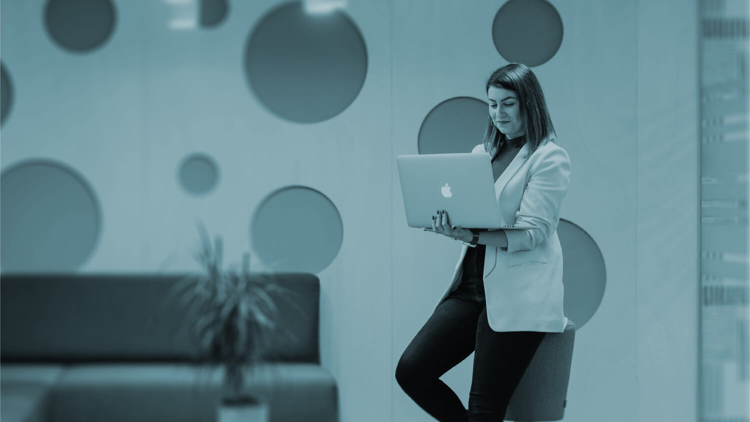 Woman standing in a modern office with circular wall designs, holding a MacBook and smiling while working, representing user-centered digital product development.