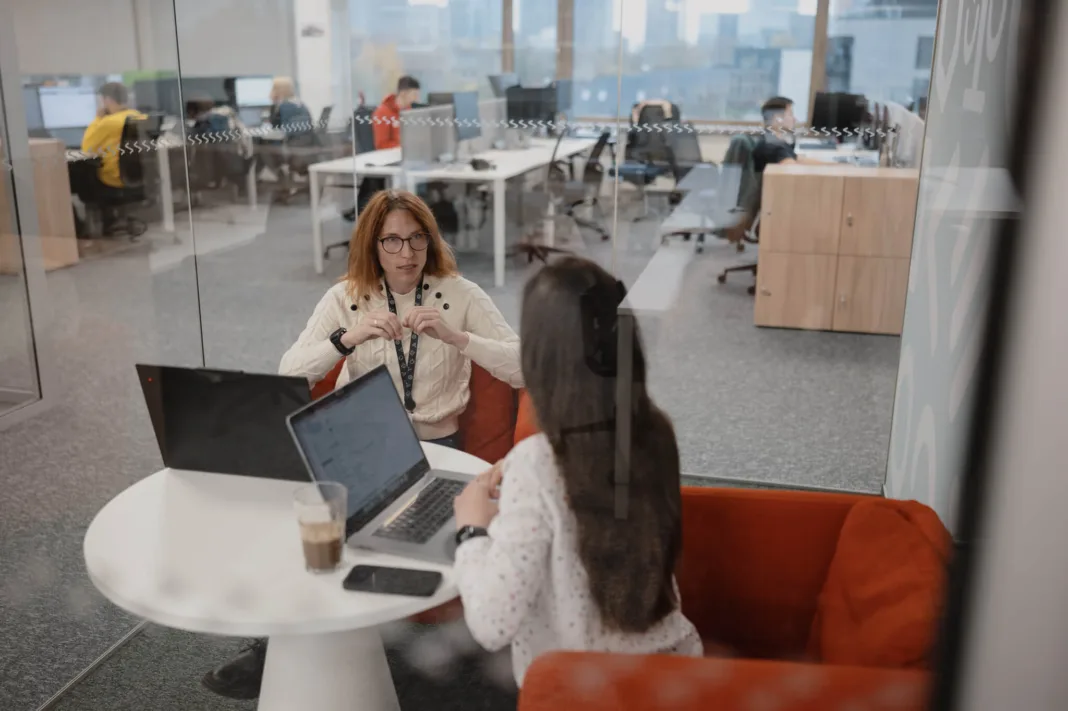 Two female colleagues sitting in a glass meeting room within an office having a discussion.
