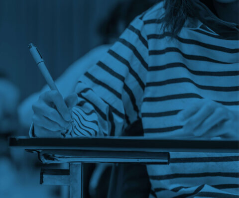 A female in a striped top sat at a desk completing an exam with a pen in her hand. The image has a blue overlay, giving the image a blue tint.